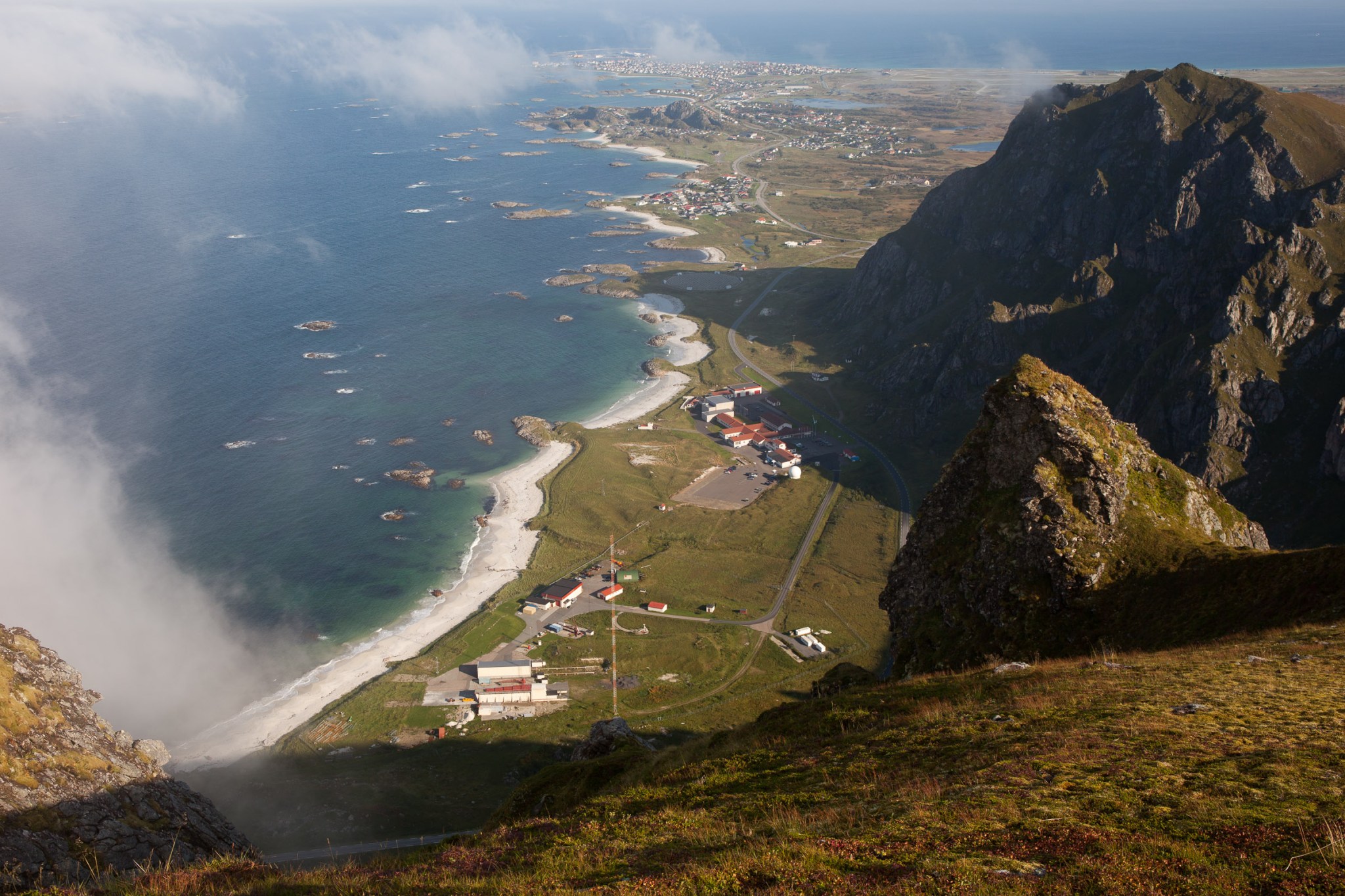 A landscape photo of a coastline in Norway, taken from the top of a mountain. Along the right side of the image is a jagged mountain leading down to a small cluster of buildings.