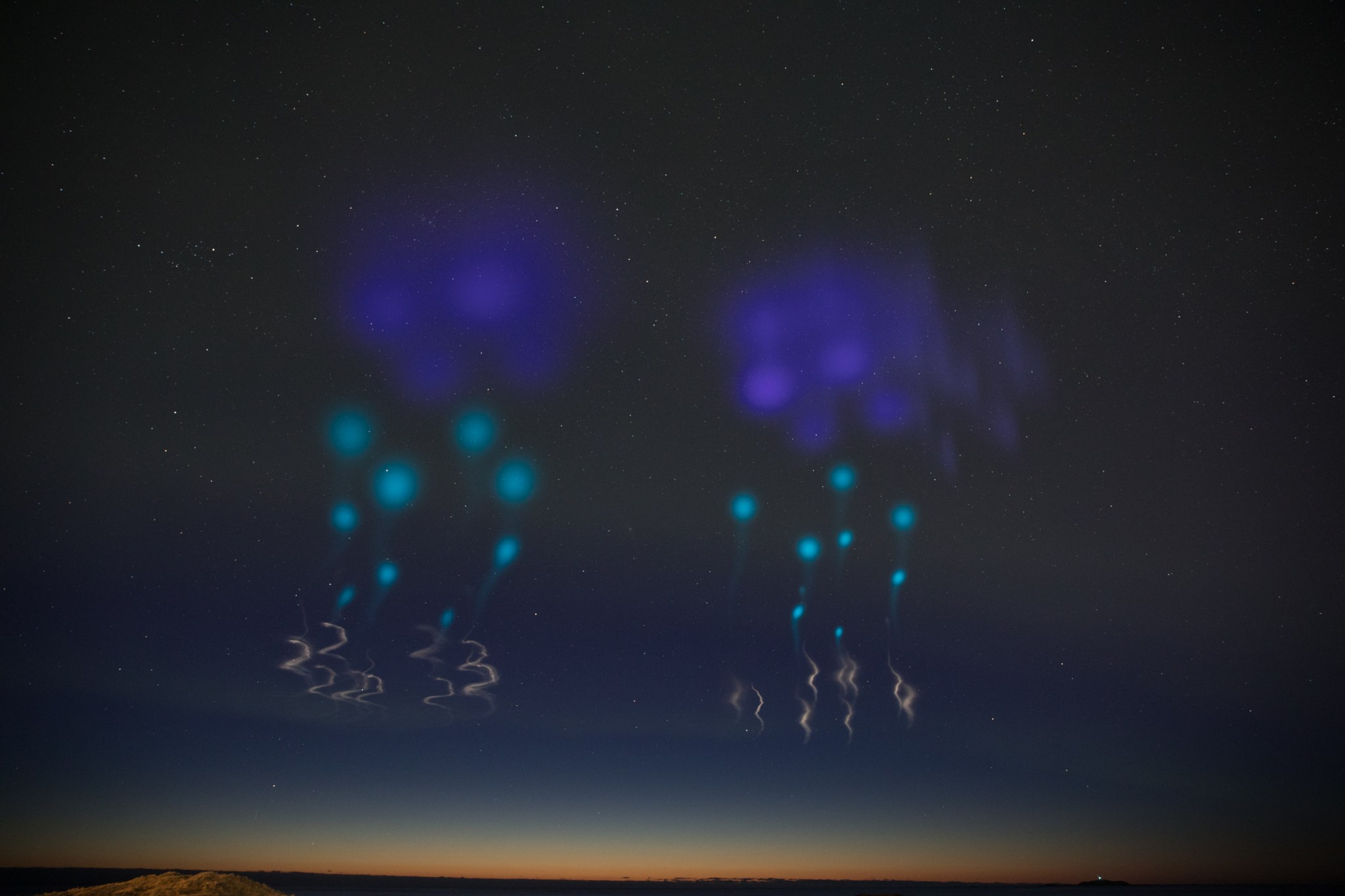 Photo of vapor tracers being released in the sky after a rocket launch. There are two sets of tracers, each has four very fuzzy deep purple spots on top. Below that are four more light blue fuzzy dots with small faded trails leading down under them, with about 3-4 smaller blue trails. Below all of that is very light orange squiggly trails leading below. The sky is clear with many small, white stars in the background, fading down into a light orange along the horizon