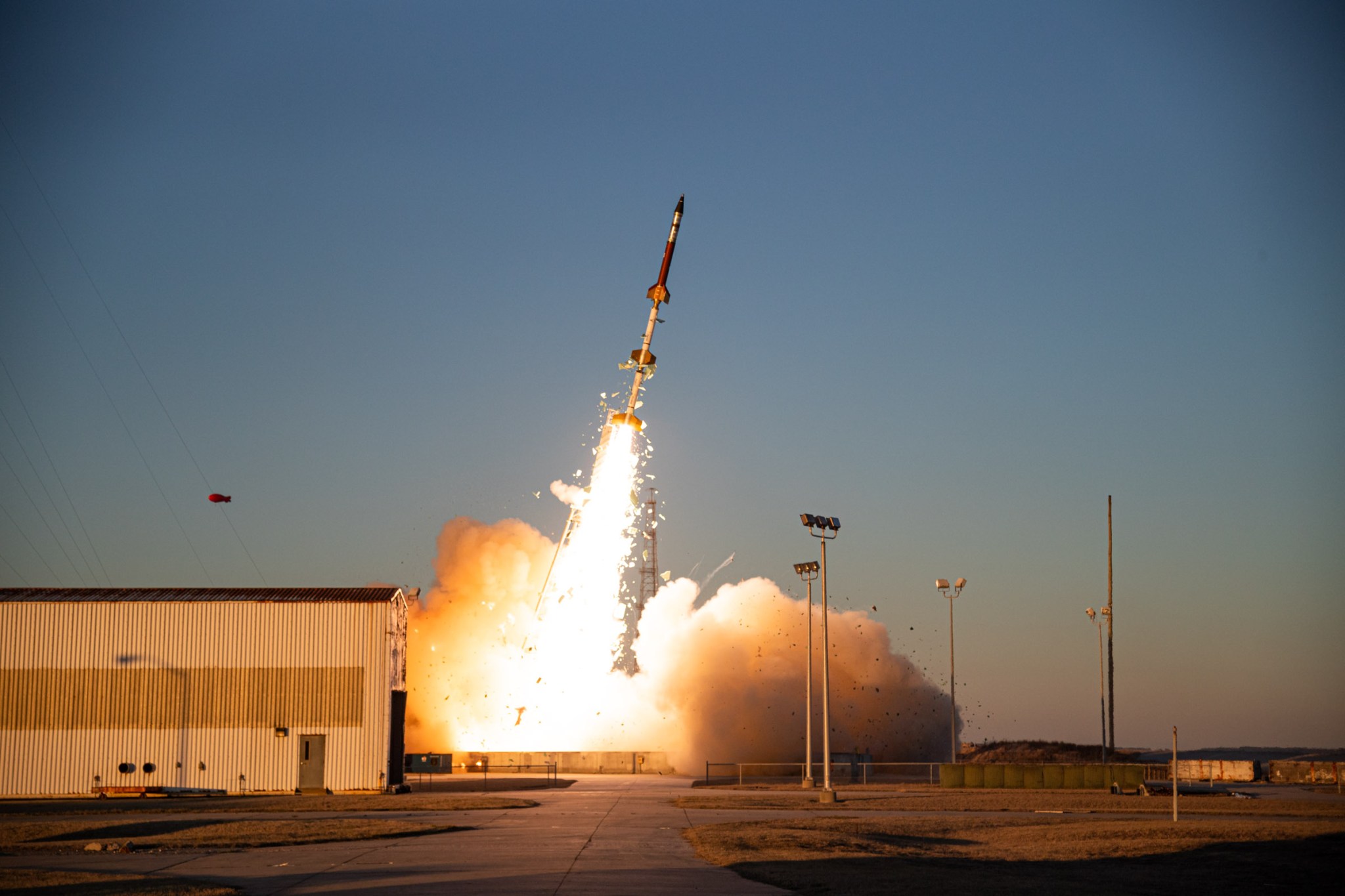 A sounding rocket seconds in mid-launch off the launch pad with a bright yellow plume underneath. The rocket, from bottom to top, has yellow fins on the bottom, a white section, bright yellow fins, a white section, bright yellow fins, a red section that leads to a silver section, and then a black cone-shaped top. In the background is a blue sky.