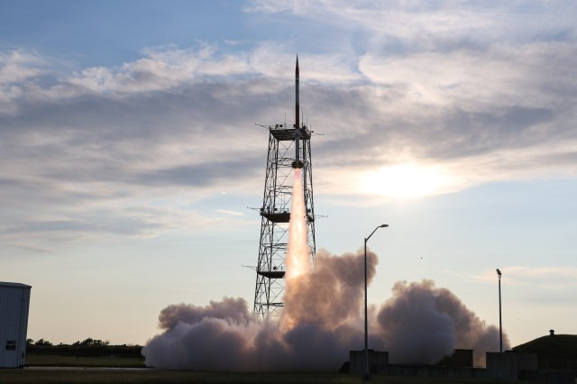 A sounding rocket seconds in mid-launch off the launch pad with a bright white plume underneath. The rocket is silhouetted by the sun behind it. In the background is a blue sky with big, puffy white clouds.