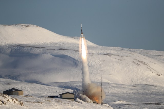 A sounding rocket launching with a plume smoke underneath against a snowy mountainside.
