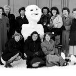 In this black and white photo, a group of women wearing heavy jackets gather around a snowman.