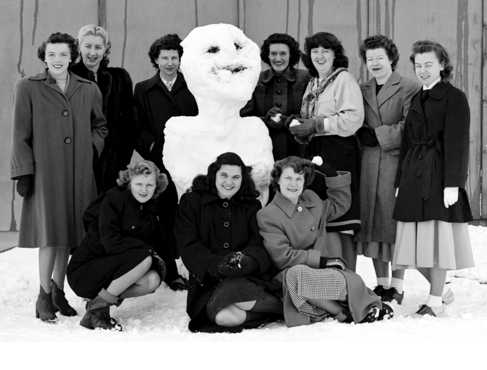 In this black and white photo, a group of women wearing heavy jackets gather around a snowman.