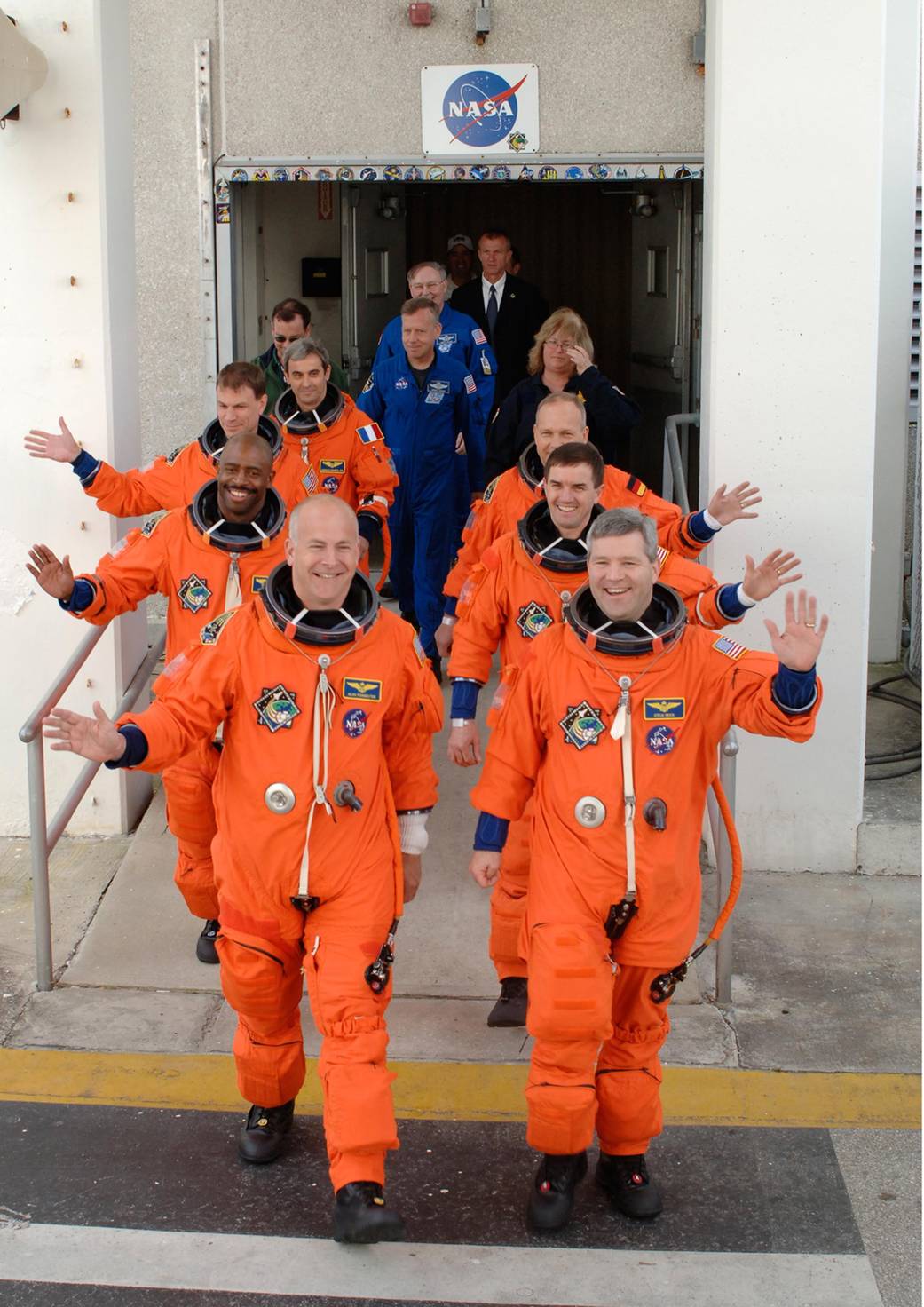 Crew members of STS-122 in orange flight suits wave as they exit the Operations and Checkout Building