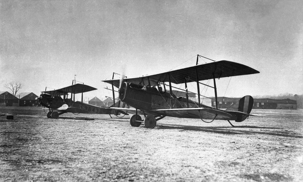 In this early photo, two Curtiss Jennies are parked on an airfield in front of hangars.