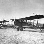 In this early photo, two Curtiss Jennies are parked on an airfield in front of hangars.