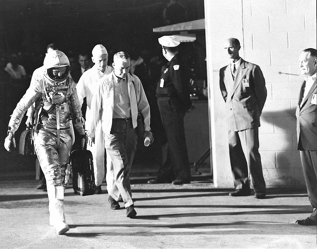 Black and white image of NASA astronaut John Glenn departs the crew quarters at Cape Canaveral Air Force Station's Hangar S. as men watch