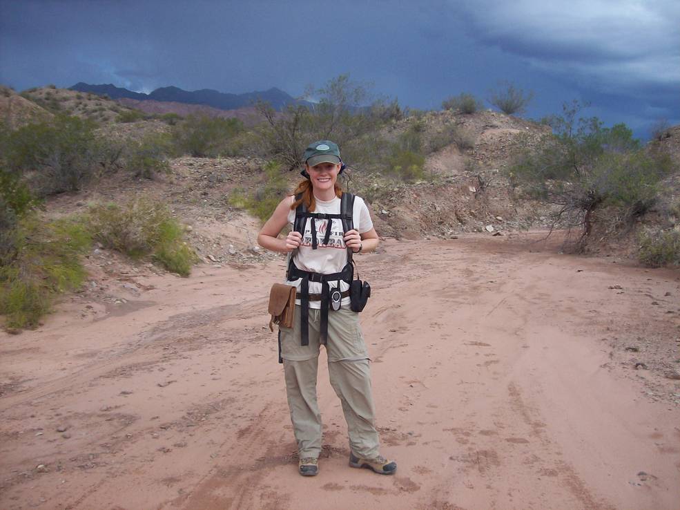 Wendy Bohon, a woman with long red hair in a ponytail, smiles and poses in front of a desert landscape. She wears a green hat, white tee, olive pants, brown boots, and a black backpack. The sky is blue-gray with clouds.