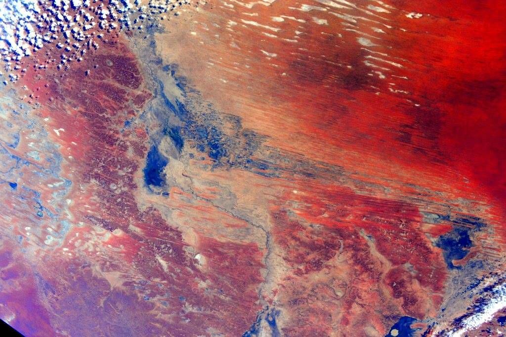 From space, image of brightly multicolored landforms with red in upper right corner, white clouds at upper left