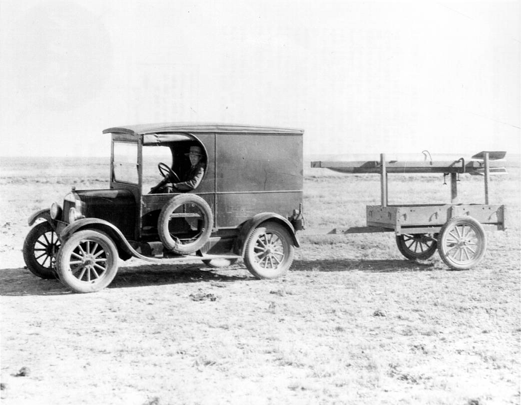 A black and white photo of a man driving an old truck towing a small rocket.