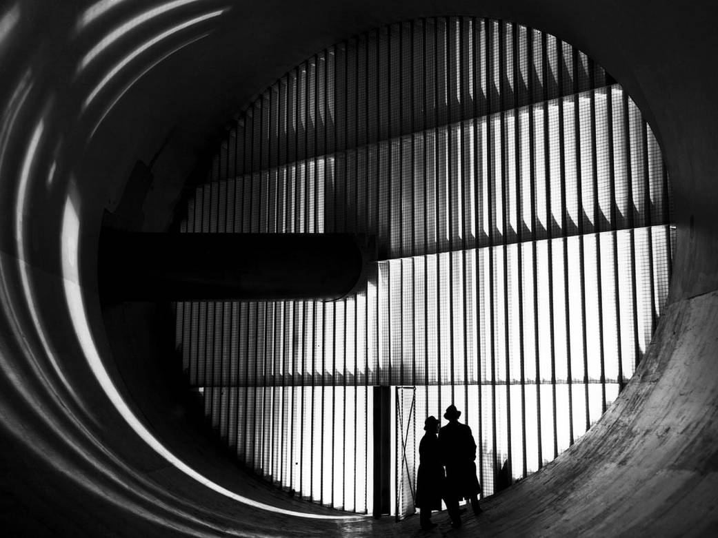 Black and white photograph of large end of wind tunnel with vertical slats running across and two people standing in front right