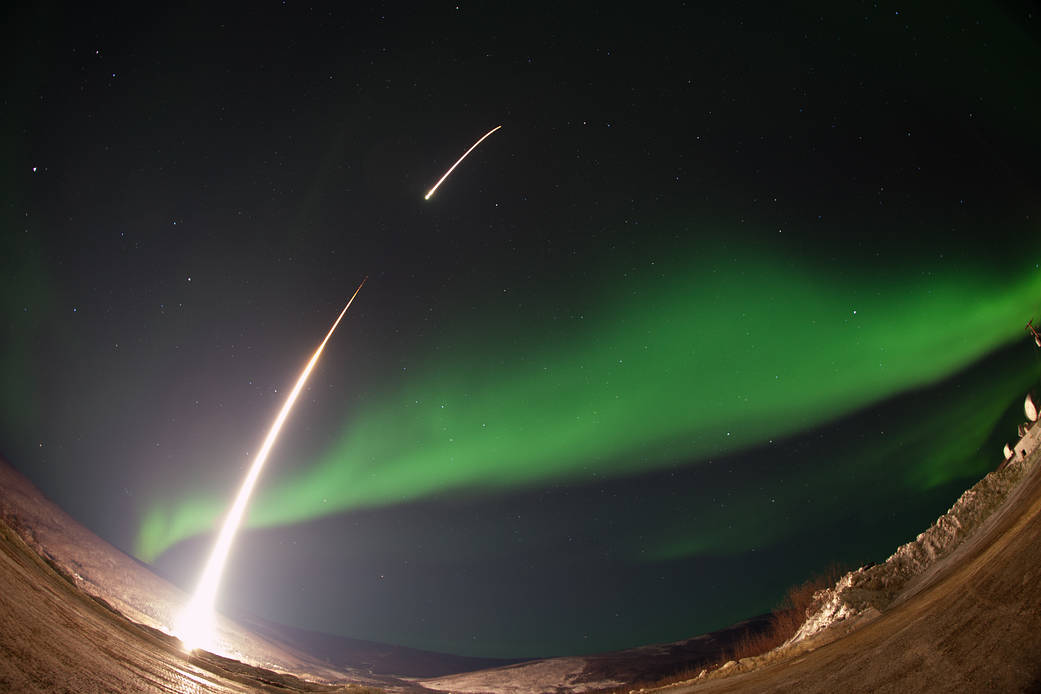 A fisheye, long-exposure photo of a sounding rocket, represented by a white streak, launching into an aurora.