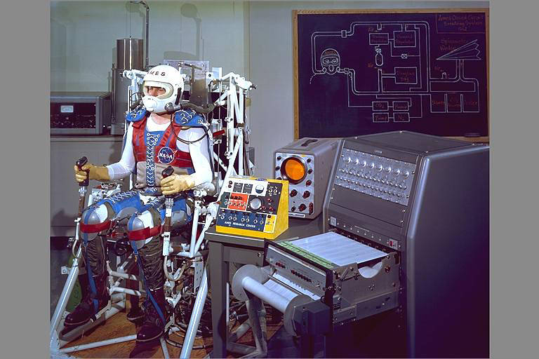 Man seated wearing mechanical system over body and helmet on head, holding brakes, with diagram of system on board at right