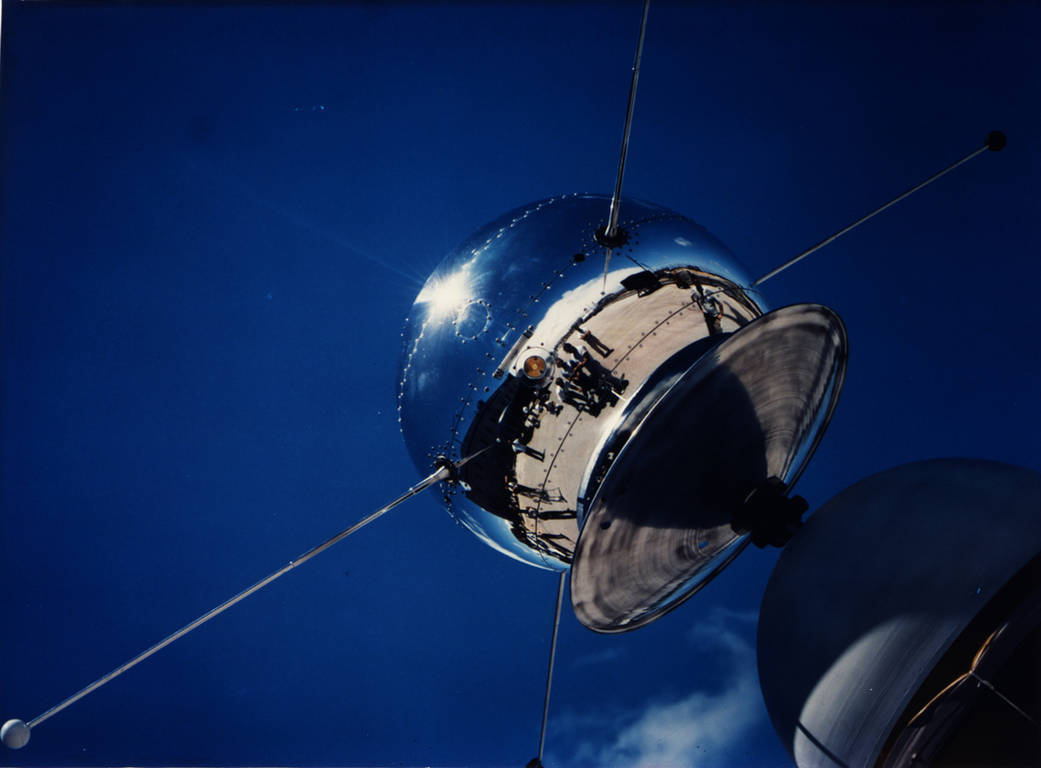 Round, silver Vanguard satellite against blue sky showing reflection of ground and personnel