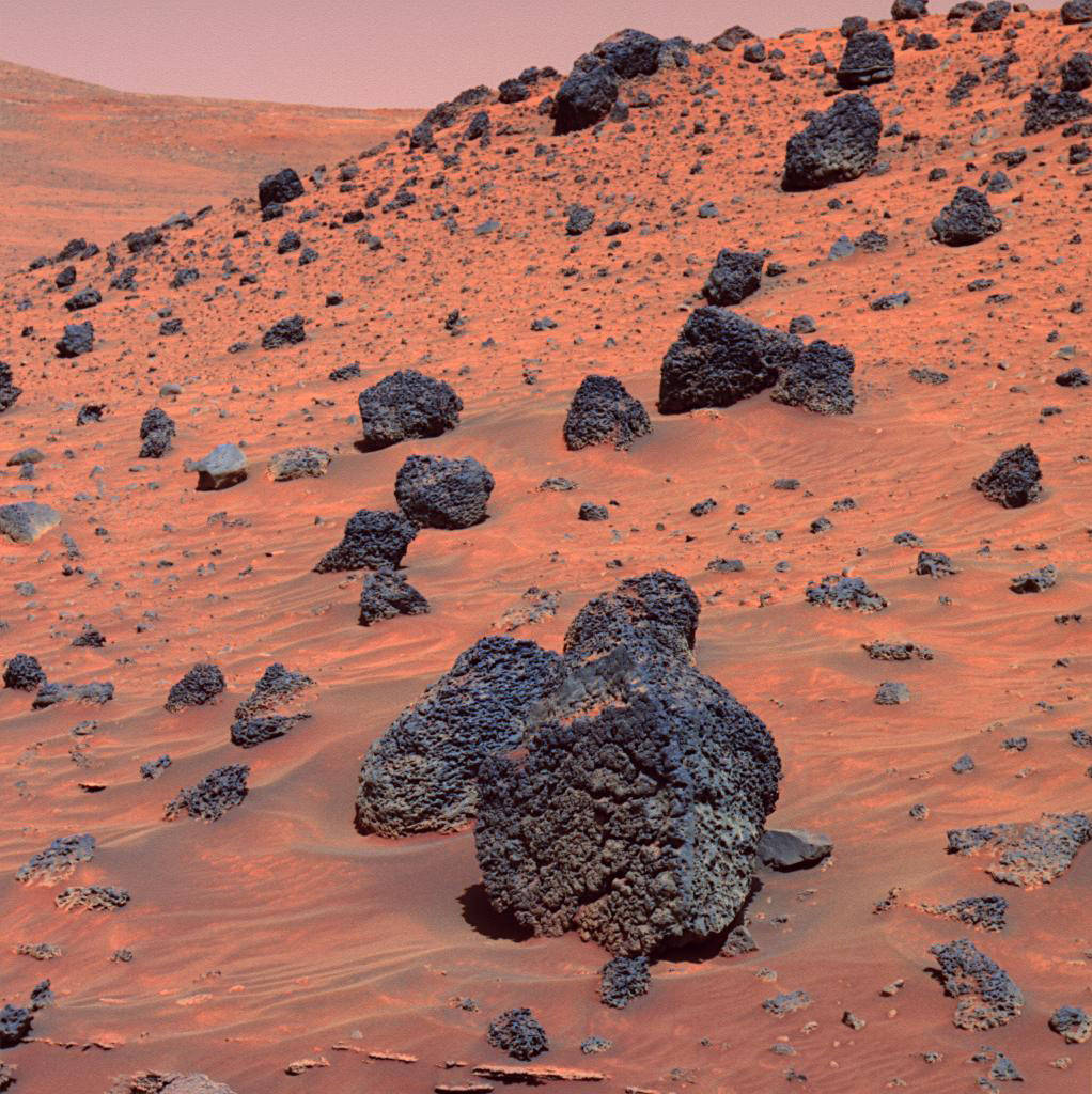 Orange red surface of Mars with large dark grey boulder in center and smaller boulders surrounding