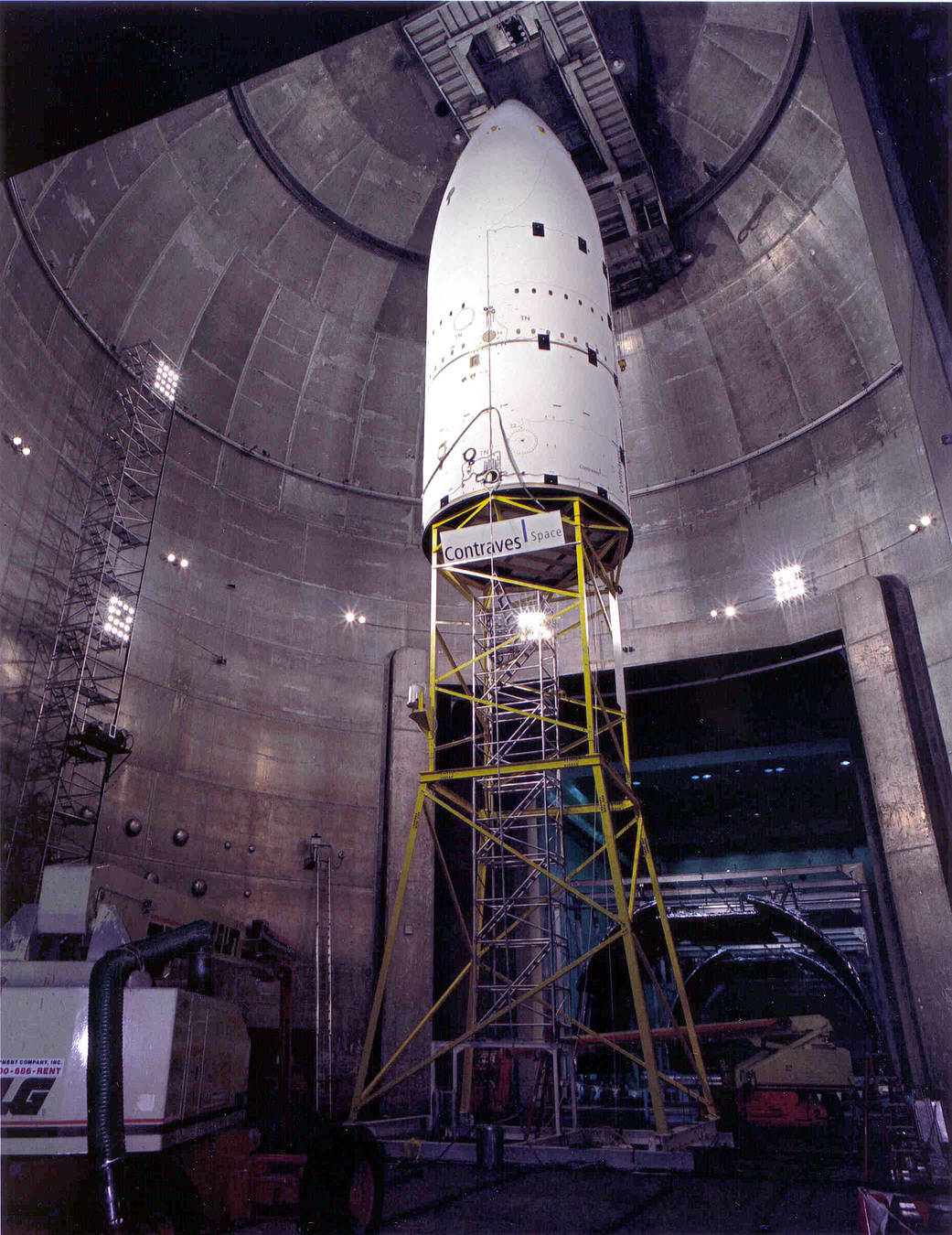 Large silver chamber with white cone shaped rocket nose on yellow scaffold in center