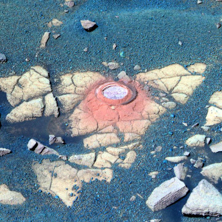 Mars surface covered in dark blue-black small rocks with flat, cracked yellow terrain in the center