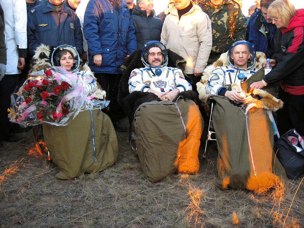 Two men and one woman in spacesuits seated with blankets after landing in Soyuz
