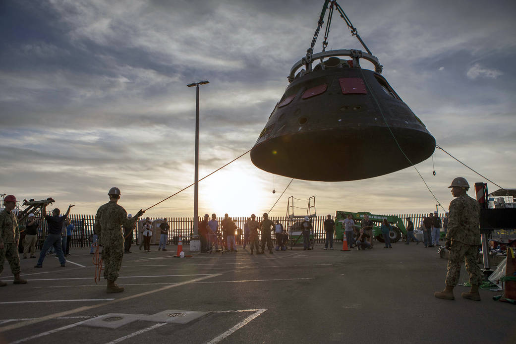 The Orion crew module is being moved by crane from its crew module recovery cradle and will be placed in the crew module transpo