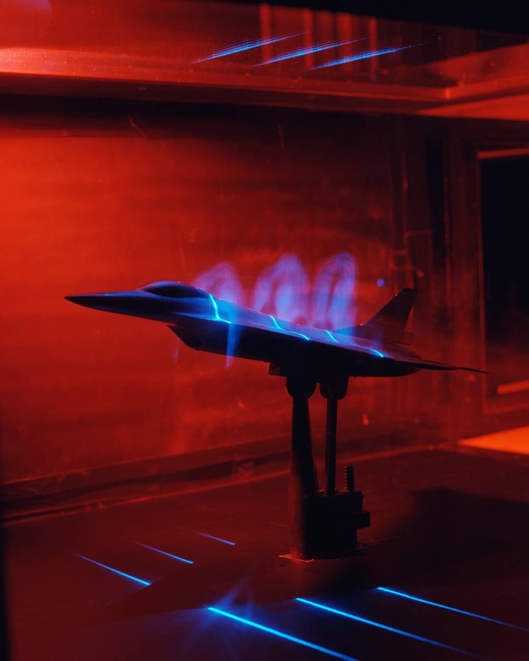 Model F-16 aircraft inside red wind tunnel with blue horizontal light beams shining from above