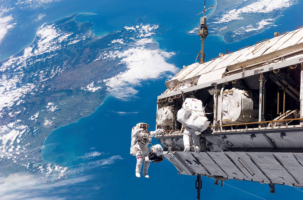 Two astronauts on spacewalk backdropped by Earth with space station module at right