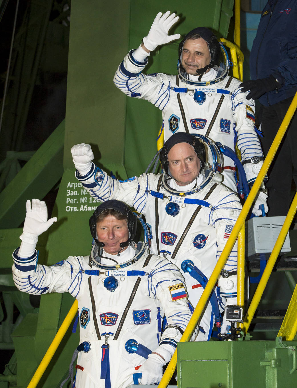 Three men in spacesuits wave as they stand on a stairway leading up to Soyuz spacecraft