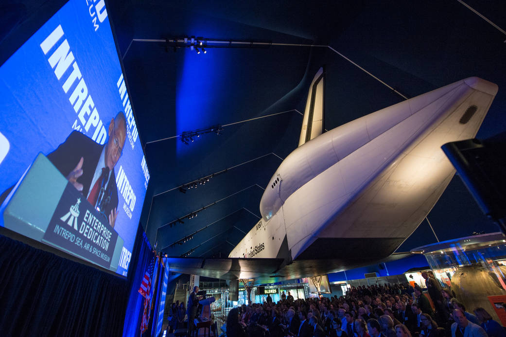 NASA Administrator Charles Bolden on screen at left speaking at ceremony with Shuttle Enterprise at right