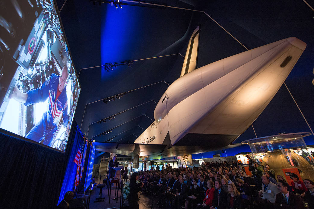 Astronaut Scott Kelly onscreen at left speaking at ceremony with Shuttle Enterprise at right