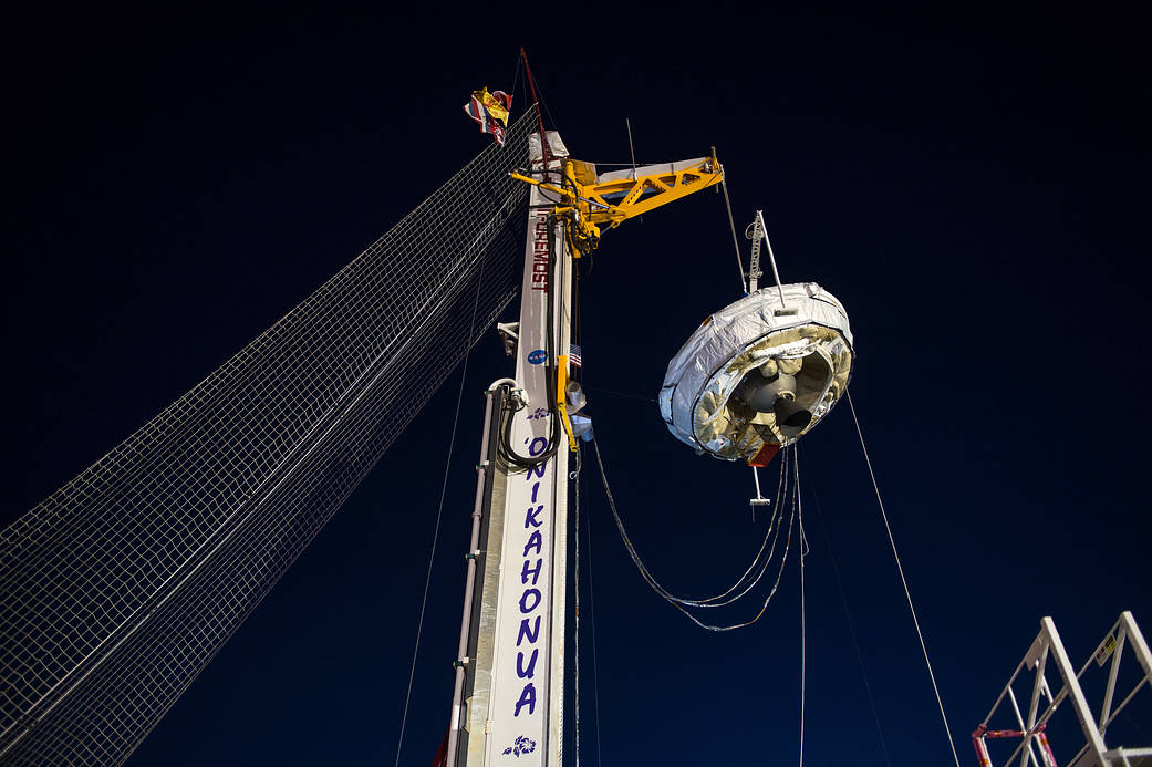 Saucer-shaped supersonic decelerator suspended from rig against night sky