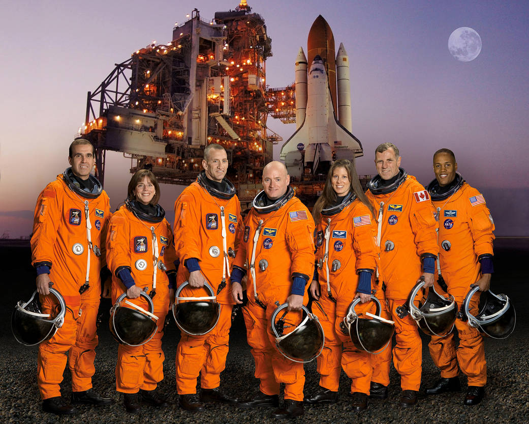Portrait of the STS-118 Crew in their orange spacesuits and holding their helmets with a backdrop of the Endeavour on the pad