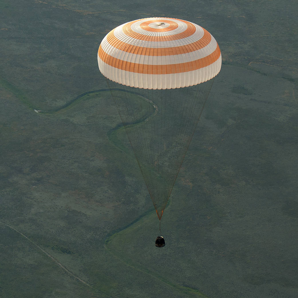 Soyuz capsule descending toward Earth with large parachute overhead