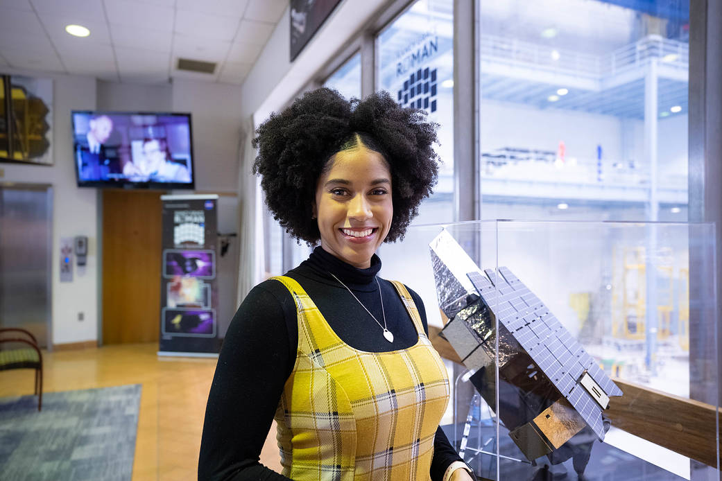 Courtney Lee, a Black American woman, looks happy and confident, smiling at the camera while standing in front of a Roman Telescope model. She's we...
