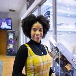 Courtney Lee, a Black American woman, looks happy and confident, smiling at the camera while standing in front of a Roman Telescope model. She's wearing a long sleeve black turtle neck under a yellow plaid dress. Her short, curly hair frames her face. 