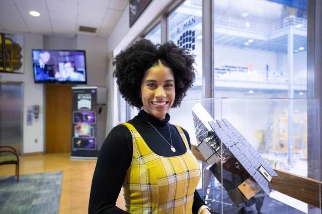Courtney Lee, a Black American woman, looks happy and confident, smiling at the camera while standing in front of a Roman Telescope model. She's wearing a long sleeve black turtle neck under a yellow plaid dress. Her short, curly hair frames her face. 