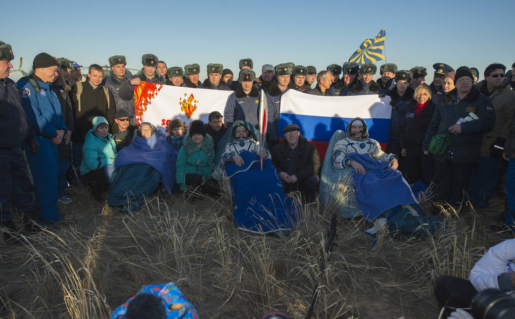 Expedition 37 landing in Kazakhstan, with crew members seated with blankets and holding Olympic torch, personnel standing nearby