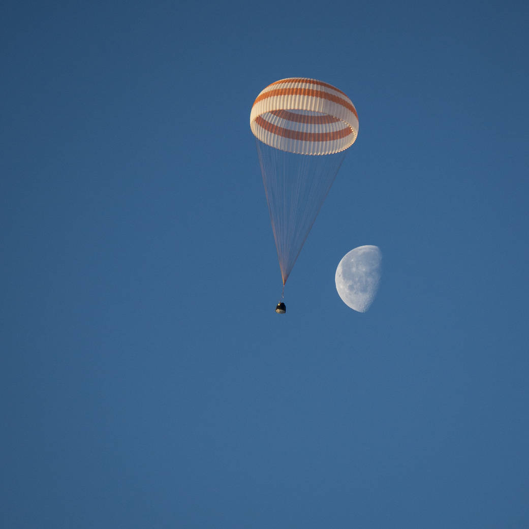 Parachute over Soyuz spacecraft against a blue sky with the moon in the background