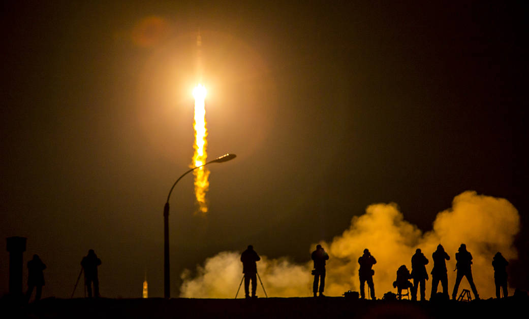 Fiery nighttime launch of Soyuz rocket at center left with media photographers in silhouette at right 