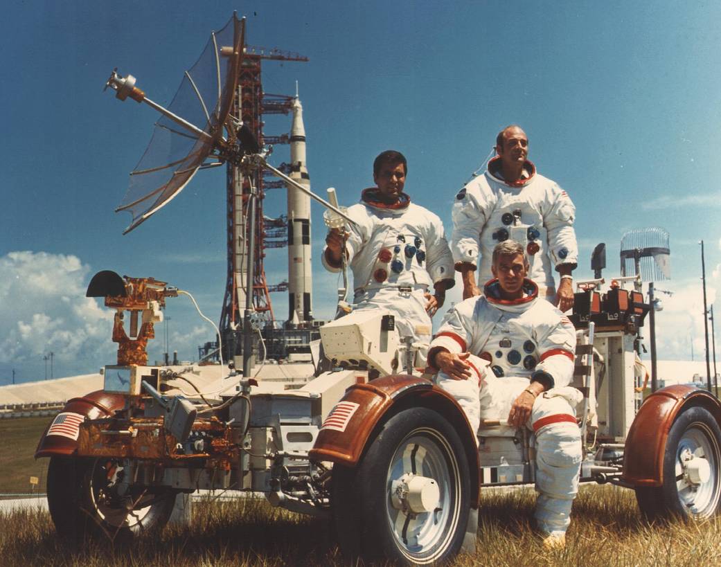 At Cape Canaveral, Apollo 17 crew in spacesuits, posing in moon rover with rocket on launchpad in background