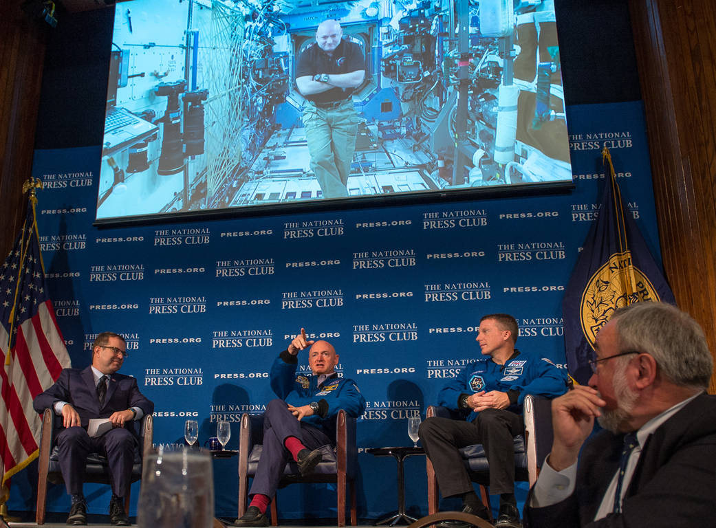 Screen showing Scott Kelly on the International Space Station and in front, seated, three men at National Press Club event