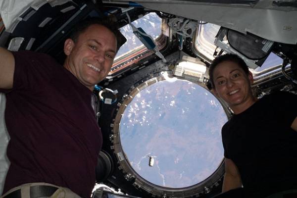 image of two astronauts posing for a photo inside the station's cupola with a capsule seen in the background as it approaches the station
