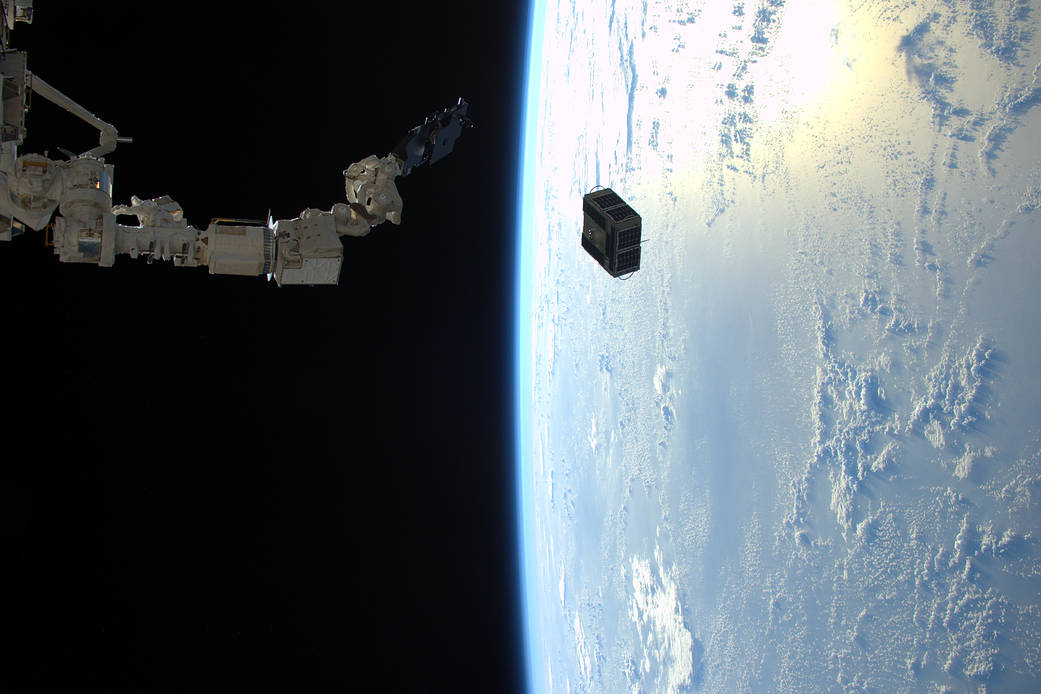 Small cube shaped satellite deployed from robotic arm with Earth visible far below