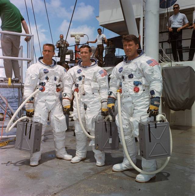 Three Apollo crewmembers in spacesuits without helmets on deck of ship