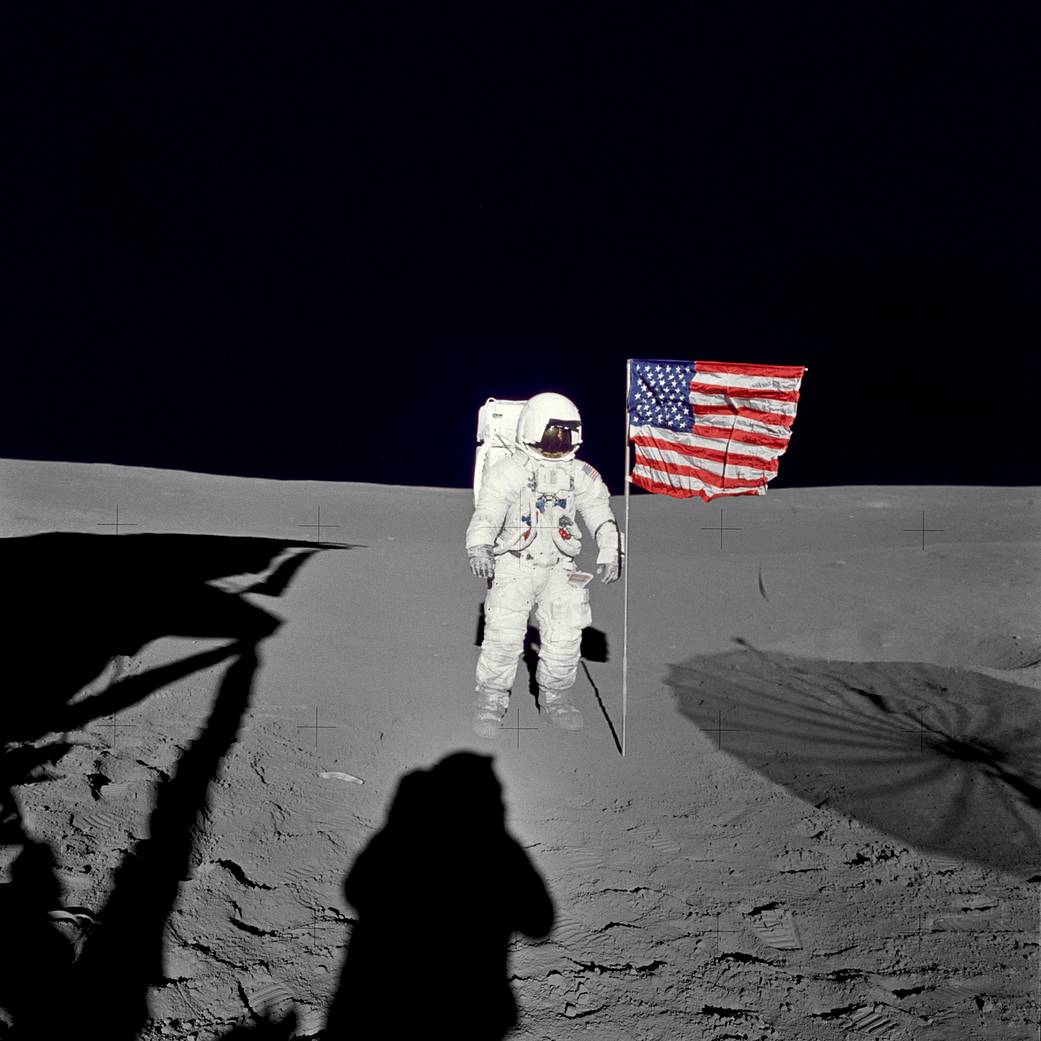 Astronaut in spacesuit standing next to U.S. flag on lunar surface with shadows in foreground