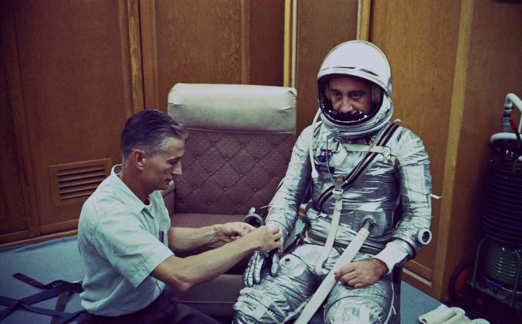 Gus Grissom wearing silver Mercury spacesuit, seated while technician checks one of the gloves