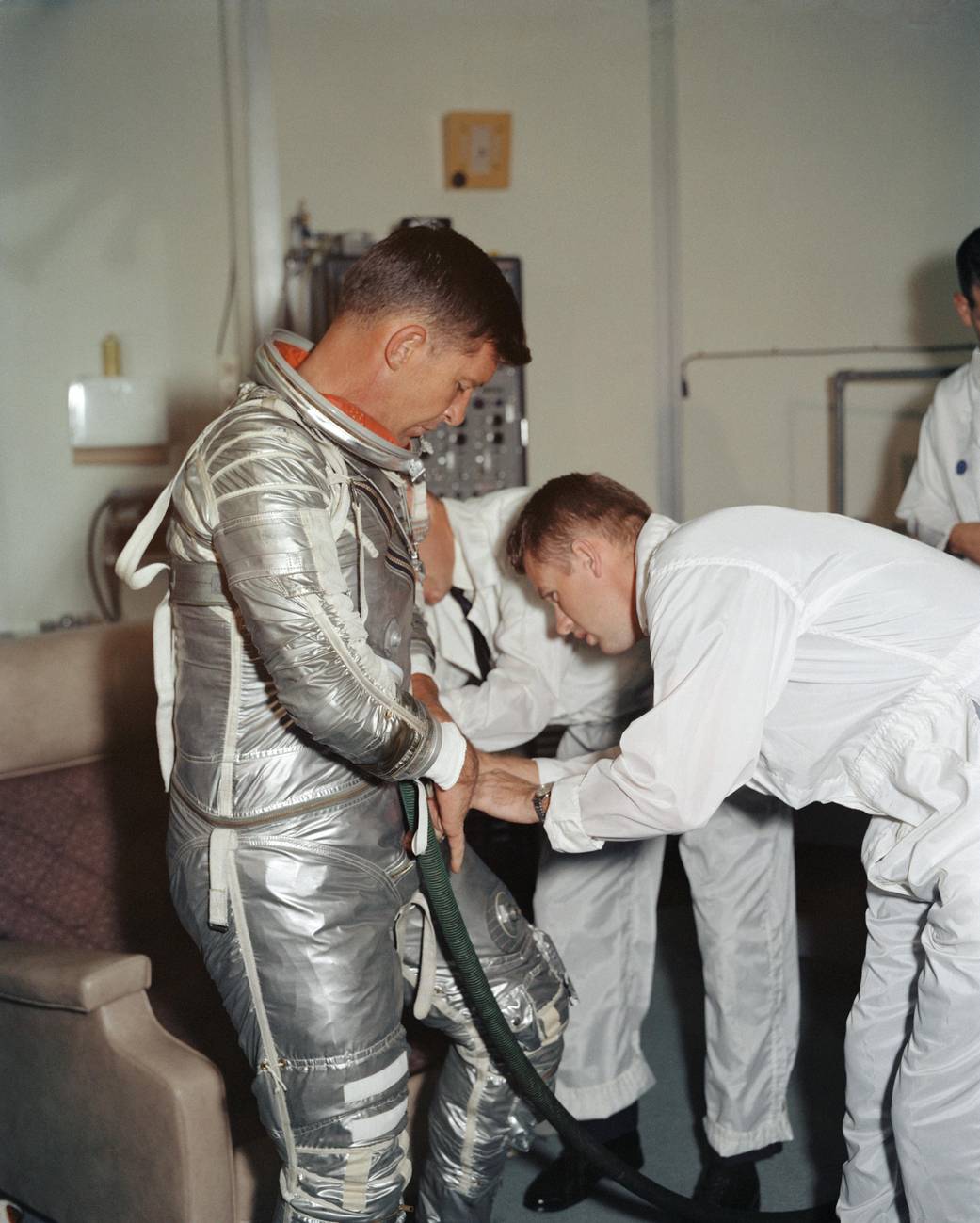 Astronaut in silver Mercury spacesuit without helmet as spacesuit is checked by technician
