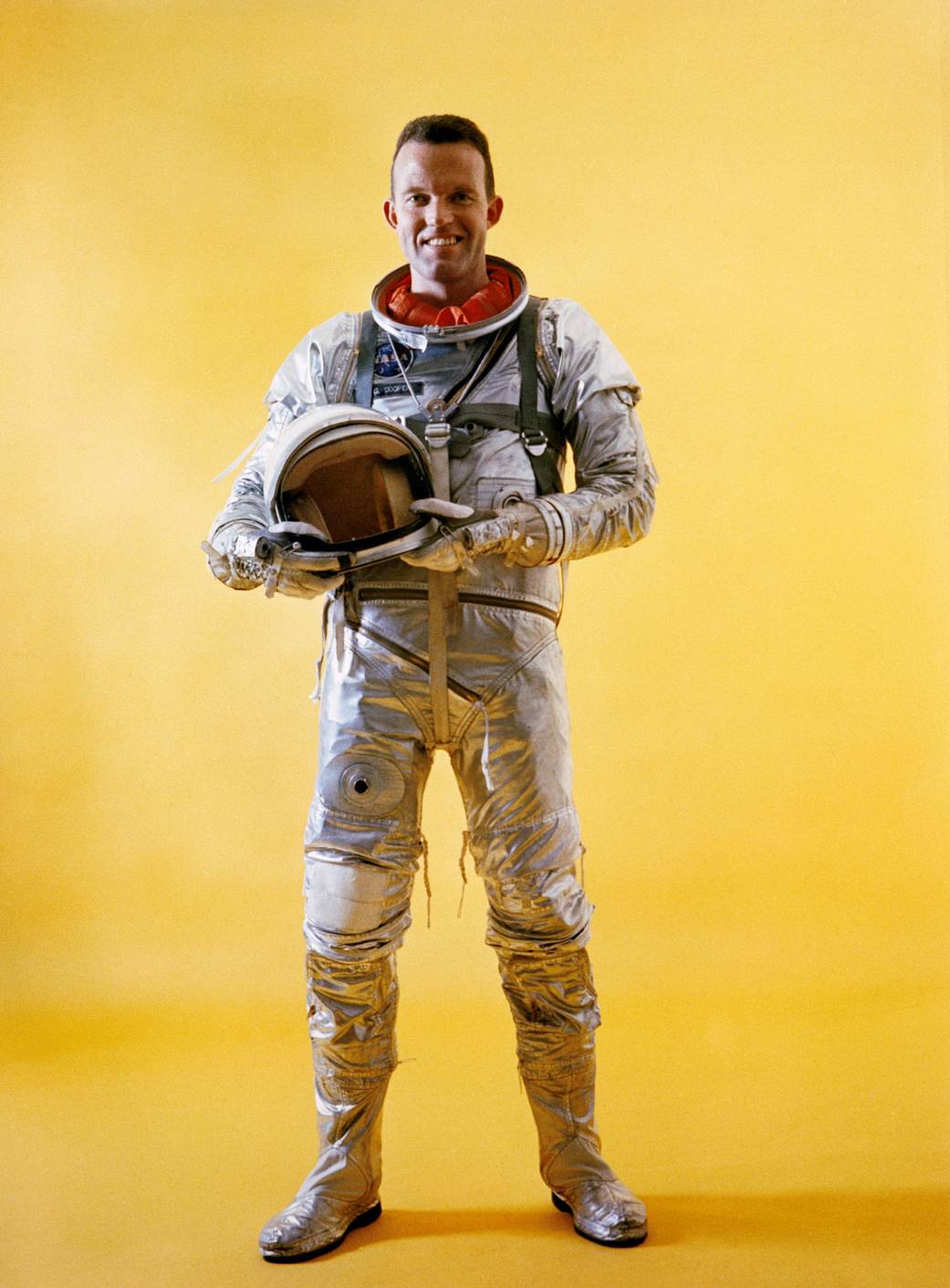 Astronaut Gordon Cooper in spacesuit holding helmet, in front of a yellow backdrop