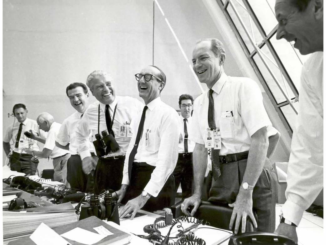 Mission control officials in white shirts and ties stand up and smile