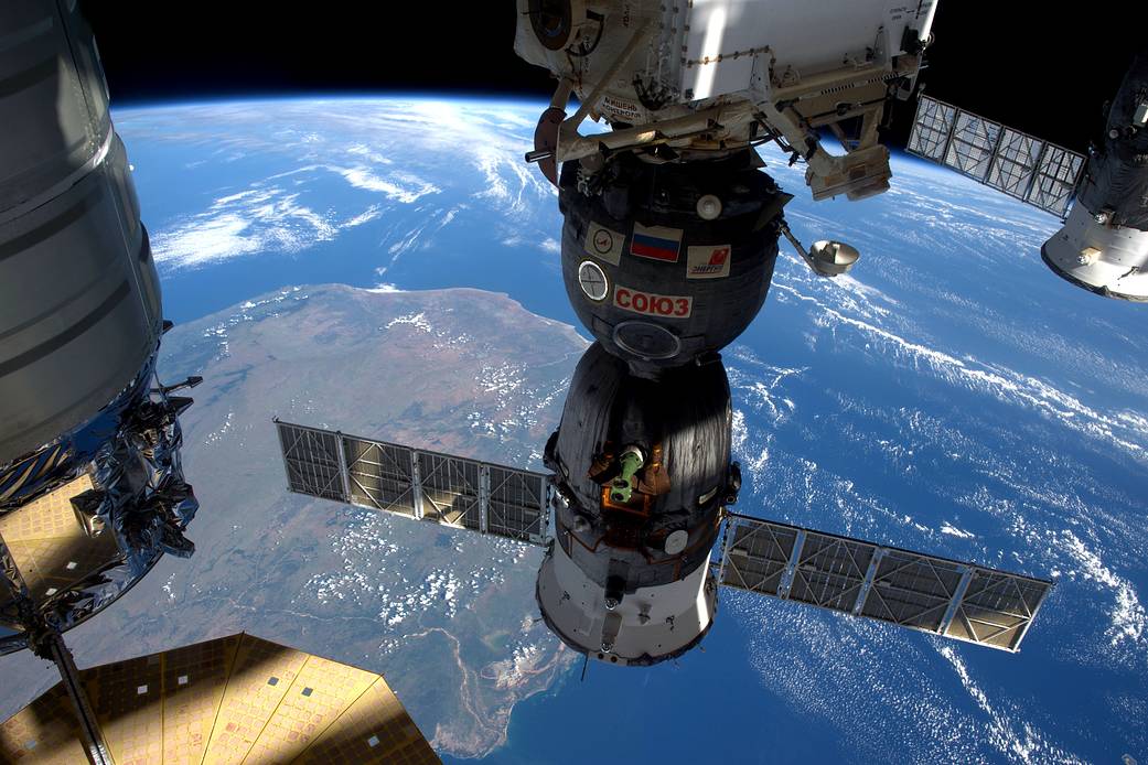 Docked Soyuz spacecraft in center of frame with Cygnus cargo craft at left and Progress craft at right with Earth below
