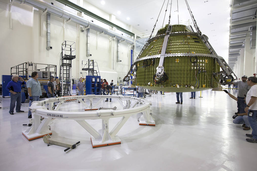 Orion crew module hoisted in air and moving toward round test stand on floor at left