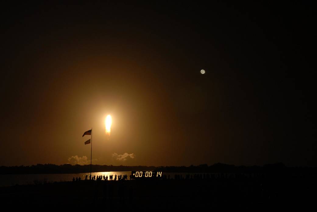 Fiery nighttime launch of Shuttle Endeavour with silhouettes of bystanders and countdown clock in foreground